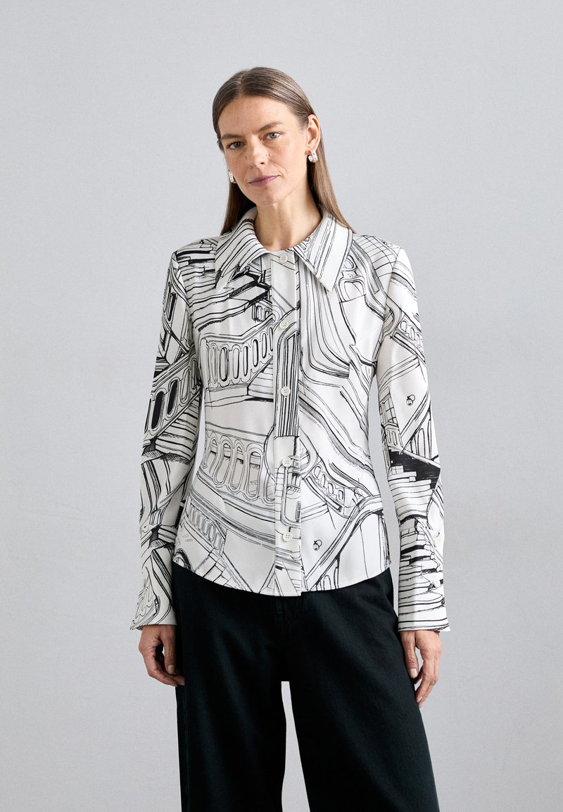 Blusa blanca con patrones lineales negros, cuello puntiagudo y cierre de botones en el frente. Mangas largas con una forma ligeramente ajustada. Combinada con pantalones oscuros.