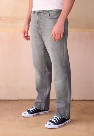 Person trägt ausgewaschene graue Jeans und schwarze Low-Top-Sneaker und steht auf einem hellbraunen Boden vor einer beigen Wand.