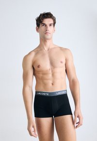Bovenlijvige man met krullend haar draagt zwarte Lacoste boxershorts, staand tegen een effen lichte achtergrond, met een lichte blik omhoog.