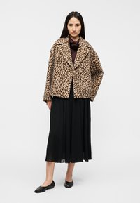 Leopard print faux fur coat with wide lapels and two front buttons, worn over a black pleated skirt. Black flat shoes complete the look.