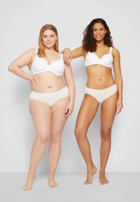 Twee vrouwen in witte bh's en beige ondergoed staan blootsvoets tegen een effen achtergrond, glimlachend met zelfvertrouwen, terwijl de een haar arm op de ander leunt.
