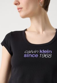 Crna pripijena majica s kratkim rukavima, sa bijelim i ljubičastim natpisom "calvin klein since 1968" sprijeda i okruglim izrezom.