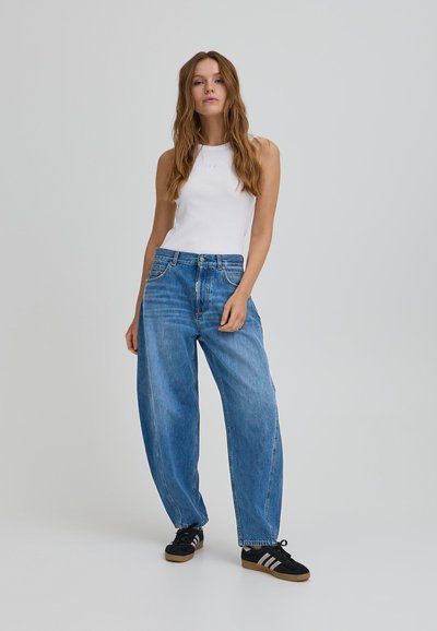 Top blanco sin mangas combinado con jeans de mezclilla azul holgados. Los jeans tienen bolsillos delanteros y cierre de botón. Zapatos negros con detalles en blanco.