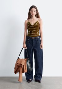 Olivengrünes Samt-Camisole mit dünnen Trägern, kombiniert mit weit geschnittenen, dunklen Denim-Jeans. Eine schwarze geflochtene Handtasche und ein orangefarbener Strickpullover sind ebenfalls enthalten.