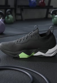 Chaussure de sport noire avec des détails verts et blancs sur la semelle, posée sur le sol de la salle de sport à proximité de kettlebells et d’une bande de résistance enroulée.