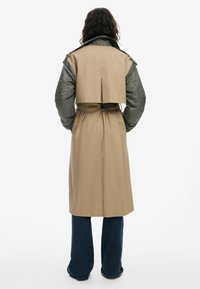 Trenchcoat met een beige ondergedeelte en een olijfgroen gewatteerd bovengedeelte, met een ceintuur in de taille en knoopaccenten.