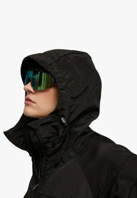 Giacca leggera nera con collo alto e cappuccio, realizzata con un tessuto strutturato. Indossata con grandi occhiali da sole riflettenti con lente verde.