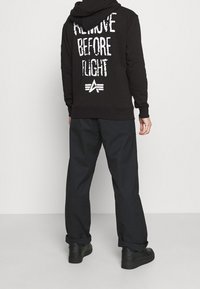 Sudadera negra con el texto blanco "REMOVE BEFORE FLIGHT" en la parte de atrás, combinada con pantalones oscuros y holgados y zapatillas negras.