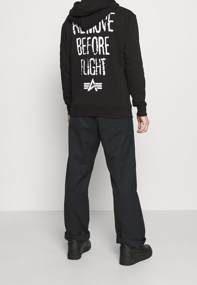 Sudadera negra con el texto blanco "REMOVE BEFORE FLIGHT" en la parte de atrás, combinada con pantalones oscuros y holgados y zapatillas negras.