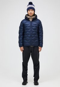 Navy puffer jacket with a quilted design, hood, and zippers; paired with black pants and a blue and white knit beanie featuring a pom-pom.