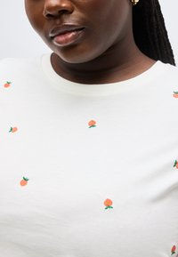 Κοντινό πλάνο ενός ατόμου που φοράει λευκό πουκάμισο με μικρή κεντημένη πορτοκαλί φρούτα και χρυσούς κρίκους σκουλαρίκια.