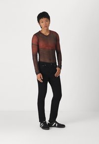 T-shirt de manga longa em rede transparente de cor castanho-escuro com gradientes vermelhos, combinada com jeans de ganga pretos e ténis pretos com riscas brancas.