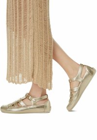 Chaussures métalliques dorées avec une semelle plate, des côtés ouverts et plusieurs lanières. Les chaussures sont associées à une robe en tricot texturé beige.