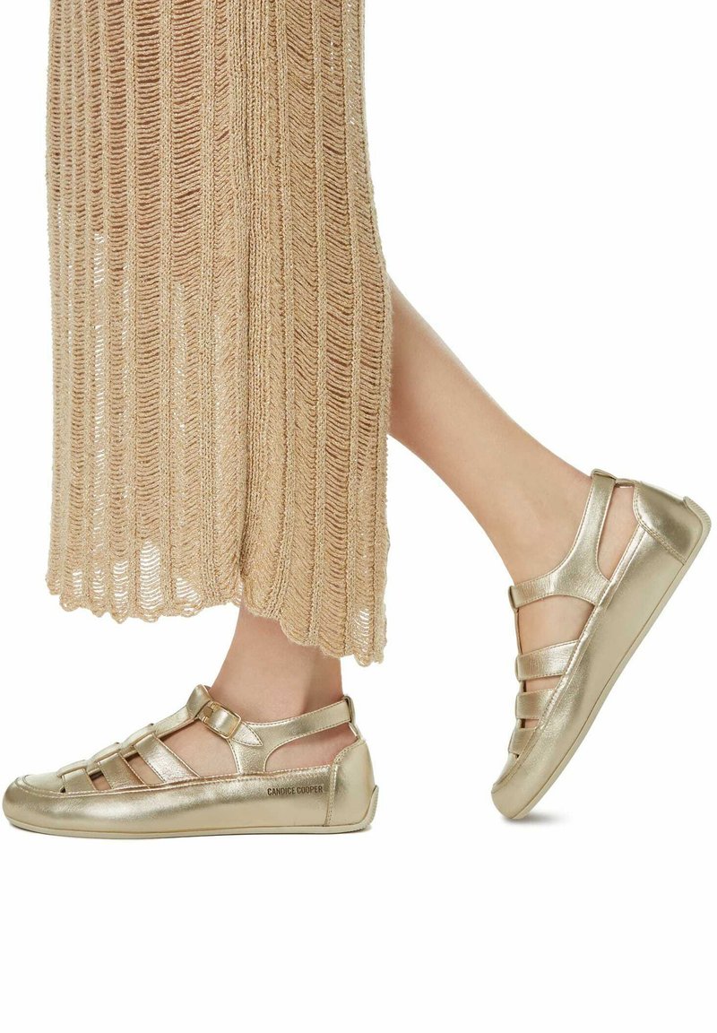 Chaussures métalliques dorées avec une semelle plate, des côtés ouverts et plusieurs lanières. Les chaussures sont associées à une robe en tricot texturé beige.