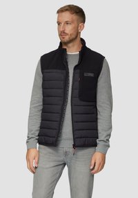 Schwarze Pufferweste mit einem Reißverschluss vorne, grauem Sweatshirt darunter und einem strukturierten oberen Bereich. Verfügt über eine Brusttasche und Logodetail.