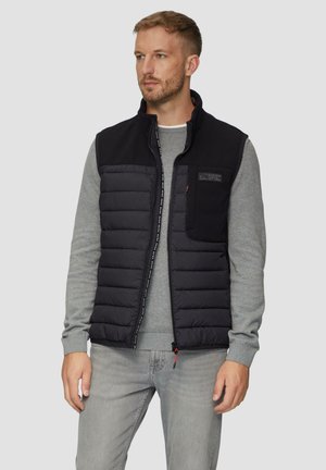 Black puffer vest with a zippered front, gray sweatshirt underneath, and a textured upper section. Features a chest pocket and logo detail.