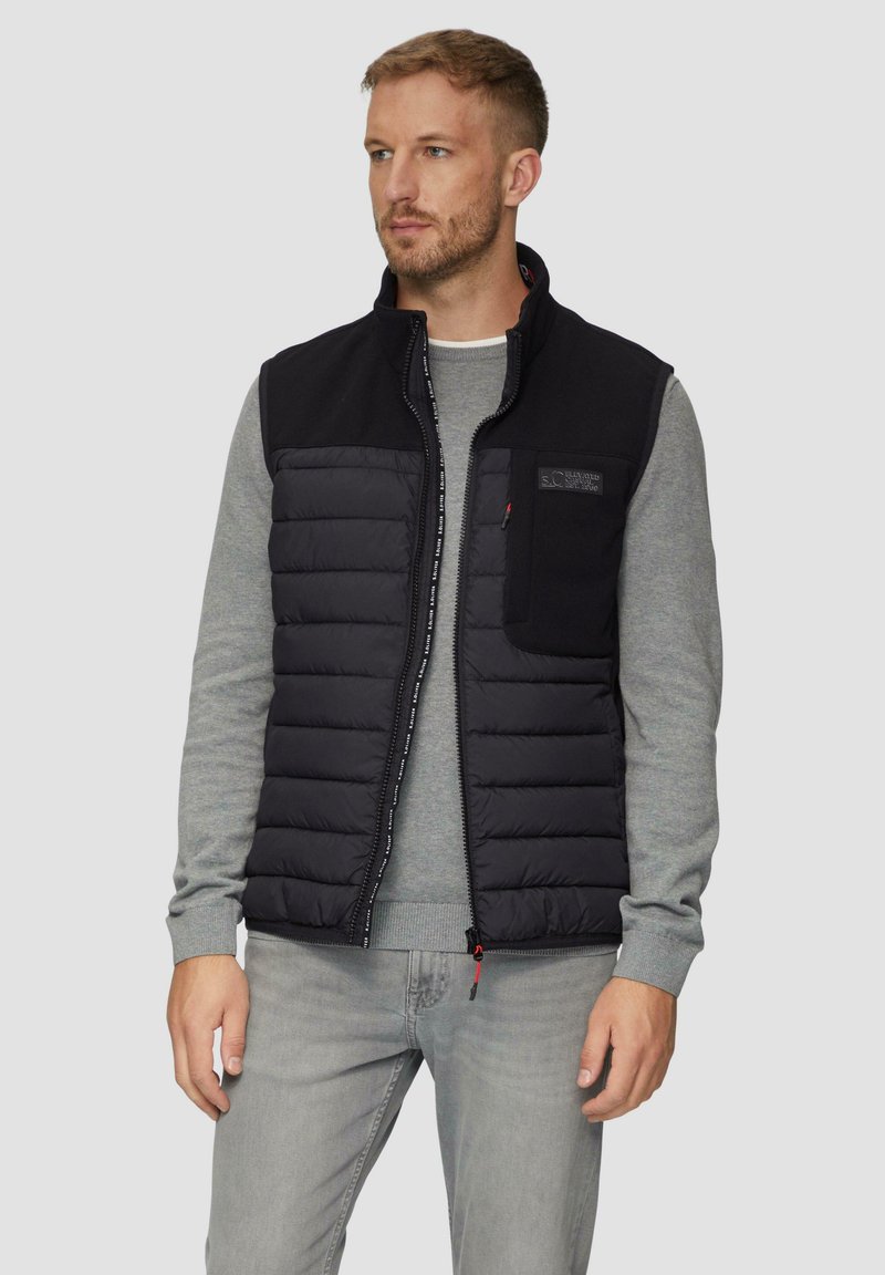 Schwarze Pufferweste mit einem Reißverschluss vorne, grauem Sweatshirt darunter und einem strukturierten oberen Bereich. Verfügt über eine Brusttasche und Logodetail.