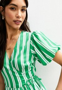 Robe enveloppante à rayures vertes et blanches avec des manches bouffantes, en tissu doux, et un détail de taille nouée. Modèle portant un simple collier en or.