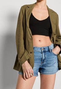 Olivově zelený lněný blazer přes černý crop top, doplněný světlými modrými denimovými šortkami. Obsahuje detaily s knoflíky a má uvolněný střih.