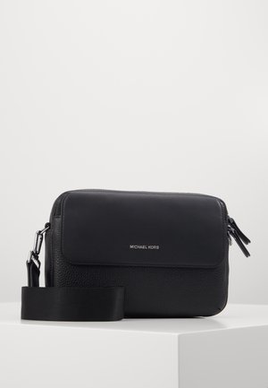 Sac bandoulière en cuir noir texturé avec rabat et logo Michael Kors argenté, sangle large réglable et double tirettes de fermeture éclair sur une surface blanche.