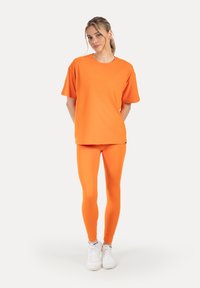 Oranje korte mouw t-shirt gecombineerd met bijpassende oranje legging, beide gemaakt van rekbare stof, aangevuld met witte sportschoenen.