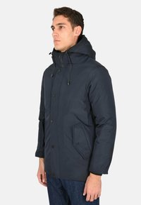 Giacca isolante blu navy con cappuccio, caratterizzata da bottoni a pressione, tasche laterali e una texture liscia. Adatta per il clima freddo.