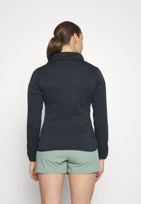 Mørkeblå fleecejakke med høj krave og tætsiddende design, der har lange ærmer og et struktureret stof, parret med lysegrønne shorts.