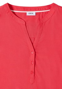 Blouse rose corail avec un col rond, trois boutons corail sur la patte de boutonnage avant et une étiquette blanche à l'intérieur du col indiquant CECIL.
