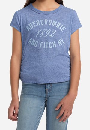 Kind trägt ein blaues T-Shirt mit kurzen Ärmeln mit dem Aufdruck „Abercrombie 1892 and Fitch NY“ und hellblaue Jeans.