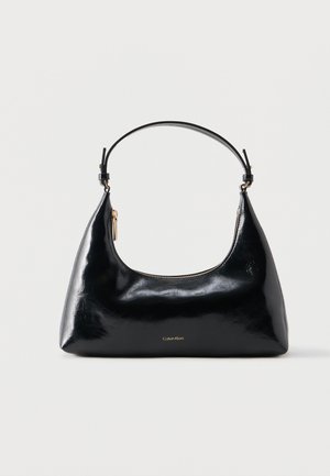 Zwarte glanzende Calvin Klein handtas met gouden rits en korte band tegen een effen witte achtergrond.