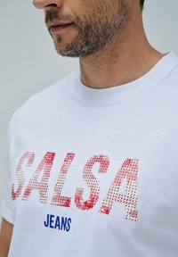 Hvid bomulds-t-shirt med stort tekst "SALSA" i rød og tekstureret mønster, samt "JEANS" i blå, rund halsudskæring, tæt på billede.