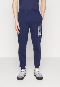 Marinblå sweatpants med elastisk midjeband och mudd. Har en vit "EA"-logotyp och texten "EMPORIO ARMANI". Mjuk tygtextur för komfort.