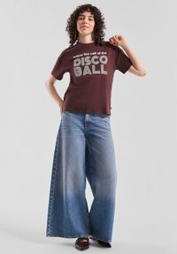 Bordeauxrotes T-Shirt mit kurzen Ärmeln und dem Aufdruck "follow the call of the DISCO BALL" in weiß. Hellblaue, weit geschnittene Jeans. Braune Schuhe.