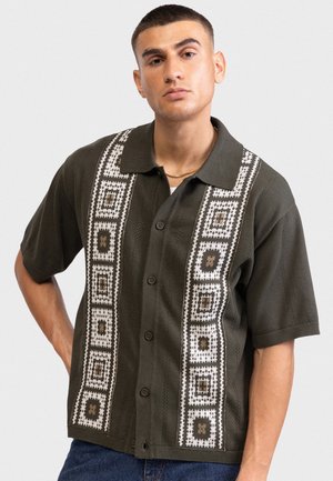 Άνδρας με κοντά μαλλιά που φοράει ένα σκοτεινό πράσινο κοντομάνικο πουκάμισο με λευκά και καφέ γεωμετρικά σχέδια και μπλε τζιν.