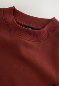 Bordowa bluza z prążkowanym, okrągłym dekoltem, gładką fakturą oraz małą metką marki widoczną wewnątrz kołnierza.