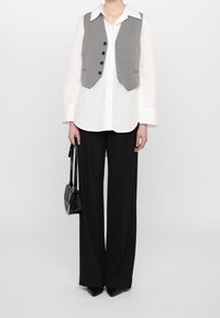 Persona que lleva una camisa blanca de manga larga, chaleco gris abotonado, pantalones anchos negros, zapatos negros puntiagudos y sostiene un pequeño bolso negro.