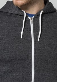 Grå dragkedja hoodie i strukturerad tyg med vit dragkedja fram och snören, samt en huva.