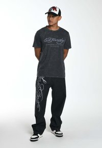 Zwarte acid-wash T-shirt met "Ed Hardy" graphic, gecombineerd met losse zwarte jeans met witte letters en zwarte sneakers met witte accenten.