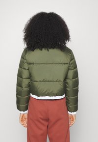 Giacca a vento verde con cuciture orizzontali, design cropped, orlo elastico bianco e polsini a coste. Indossata su pantaloni della tuta arancioni.