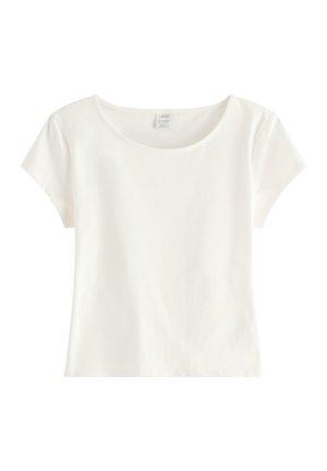 Unge kvinde står med hænderne i lommerne, iført en hvid top med korte ærmer, lyseblå satinbukser og beige broderede flade sko.