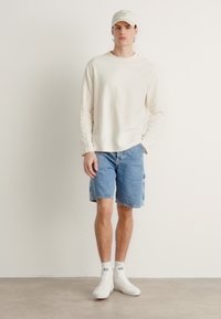 Svetlá krémová košeľa s dlhým rukávom, modré denimové cargo šortky, biele tenisky a svetlobéžová čiapka s textom. Jednoduchý, neformálny outfit.