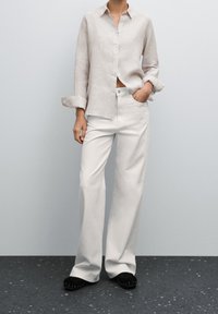 Jasno beżowa lniana koszula z zapięciem na guziki i podwiniętymi rękawami, w połączeniu z szerokimi białymi spodniami i czarnymi butami wsuwanymi.
