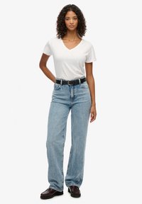 Witte v-hals t-shirt, blauwe wijde jeans, zwarte riem met zilveren gesp, bruine schoenen, model met krullend haar staat op een effen achtergrond.