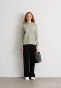 Sudadera verde claro con logo, pantalones anchos negros, zapatillas blancas y un bolso negro texturizado con patrón de zigzag, contra un fondo neutro.