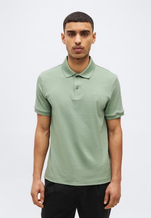 Bărbat stând cu fața înainte, purtând un tricou polo cu mâneci scurte de culoare verde deschis și pantaloni negri, pe un fundal simplu.