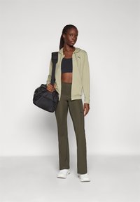 Grön zip-up hoodie, svart crop top, olivgröna leggings och vita skor. Svart träningsväska med axelrem. Enkelt, atletiskt design.