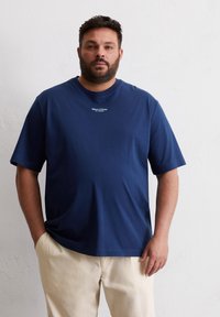 T-shirt bleu marine à manches courtes en tissu doux, avec un col rond et un logo blanc subtil sur la poitrine. Porte avec un pantalon clair.