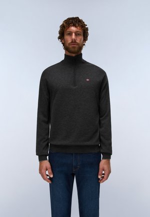 Mann mit lockigen Haaren und Bart trägt einen dunkelgrauen Quarter-Zip-Pullover und blaue Jeans, blickt nach vorne vor einem schlichten, hellen Hintergrund.