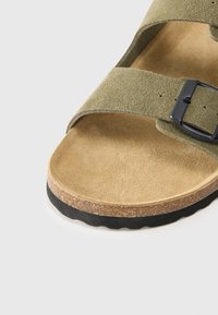 Sandale en suède vert olive avec une boucle noire, semelle intérieure rembourrée beige et semelle texturée marron, présentant un design de sangle simple.