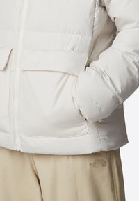 Vit puffjacka med dragkedja, stora framfickor och broderad logotypdetalj; mjuk, slät textur och avslappnad passform.